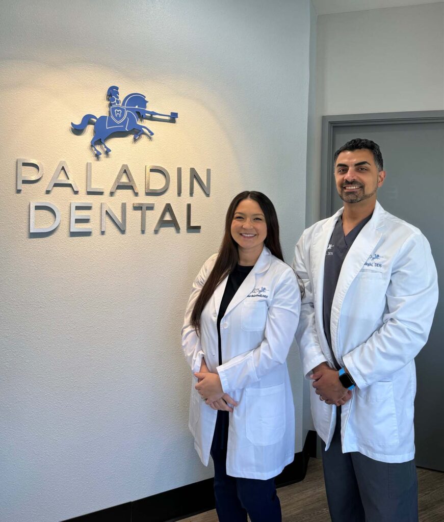 Dr. Sadeghi and Dr. Beladinelli smiling confidently in front of the Paladin Dental logo at their clinic in Fresno, CA. They are dressed in professional white lab coats, standing against a wall that displays the clinic's logo, which includes a stylized blue knight on horseback above the bold lettering of 'PALADIN DENTAL'. Their friendly demeanor and professional attire reflect the welcoming and expert care they provide.