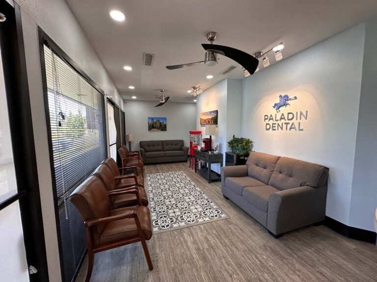 Spacious and modern waiting area at Paladin Dental in Fresno, CA. The lobby features comfortable seating arrangements with a mix of leather armchairs and a plush gray sofa, complemented by a decorative area rug. The office's logo is illuminated on a light blue wall, adding a touch of elegance. Large windows with blinds allow natural light to fill the room, enhancing the welcoming atmosphere.