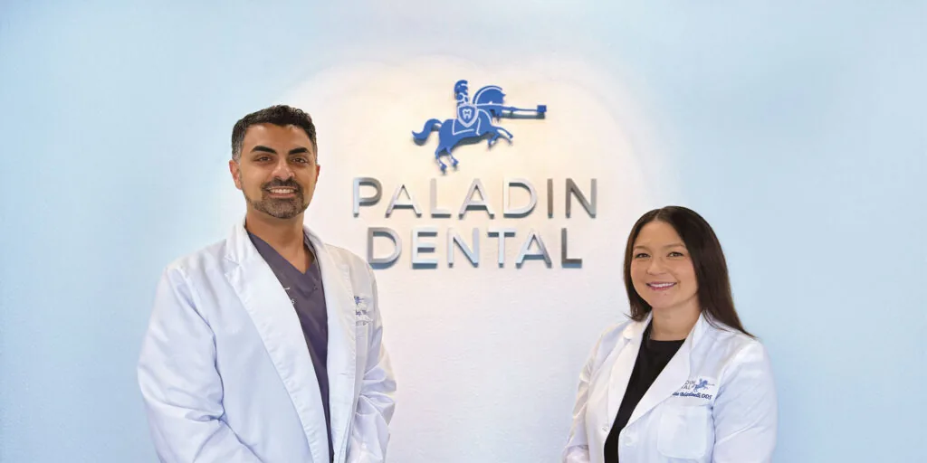 Two individuals wearing white lab coats standing in front of a wall with the text 'PALADIN DENTAL' and a knight on a horse logo.